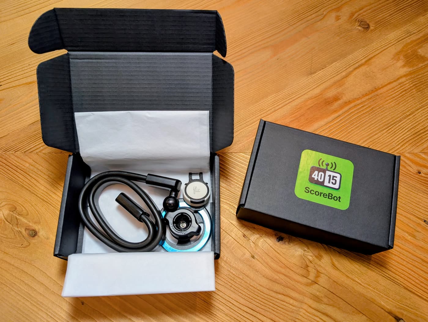A small black shipping box open to show the ScoreBot button, clip and phone mount wrapped in tissue paper, next to a closed box with the ScoreBot logo on the lid